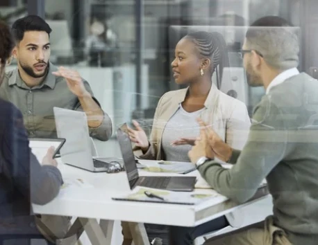 People discussing ideas around a table with laptops in a glass-walled workspace